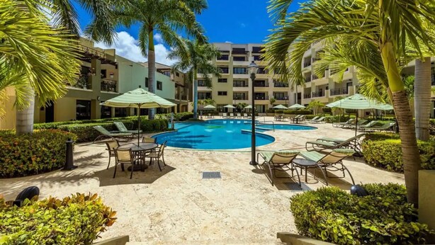 Área de piscina em resort, com espreguiçadeiras e guarda-sóis, cercada por palmeiras e um edifício de vários andares.