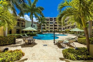 Zona de piscina en un resort con sillones de descanso y sombrillas, rodeada de palmeras y un edificio de varios pisos.