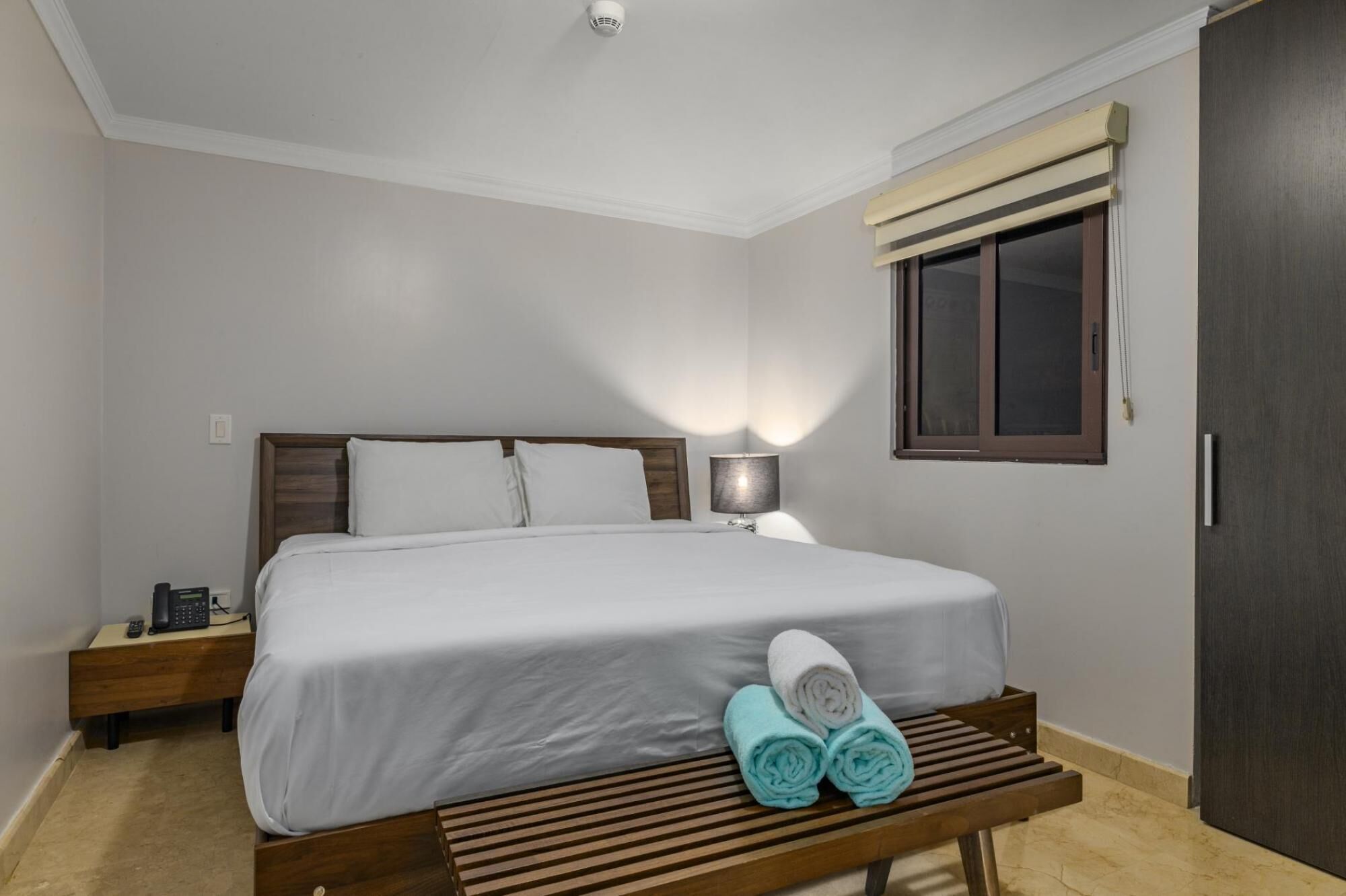 A hotel room with a bed, bedside table, lamp, and a bench with towels.