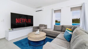 A modern living room with a large flat-screen TV displaying 'NETFLIX', a grey sofa, a round coffee table, and a view of a swimming pool through large windows. - Boggey s Villas - Xander Bogaerts 5BR 7BA w Pool (San Nicholas Noord)