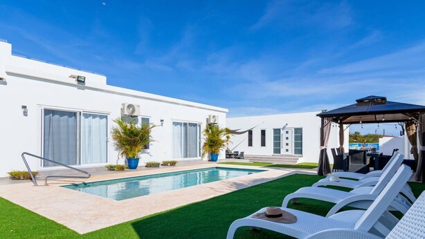 A modern outdoor pool area with a clear blue sky, a white building with large windows, and a covered seating area with lounge chairs. - Boggey s Villas - Xander Bogaerts 5BR 7BA w Pool (San Nicholas Noord)