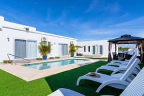 A modern outdoor pool area with a clear blue sky, a white building with large windows, and a covered seating area with lounge chairs. - Boggey s Villas - Xander Bogaerts 5BR 7BA w Pool (San Nicholas Noord)