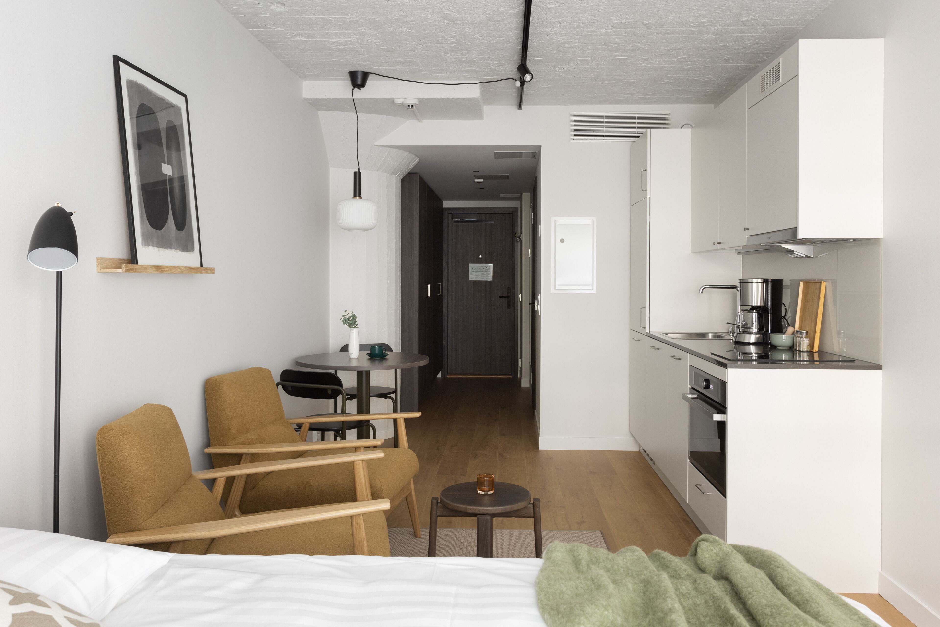A compact hotel room with a bed, a sofa, a small table, a desk, and a kitchenette.