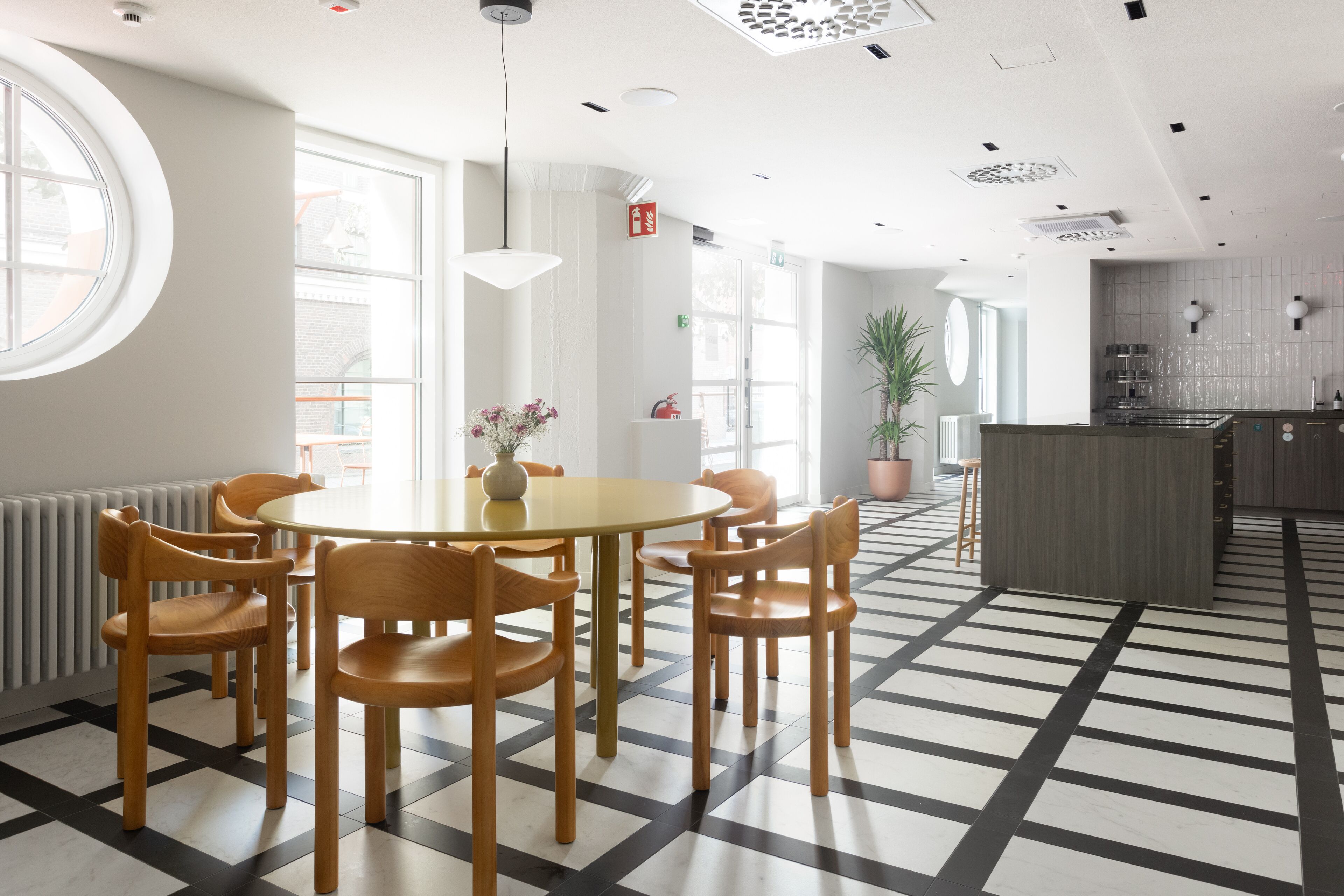 Uma sala de jantar moderna com uma mesa redonda e cadeiras de madeira, uma planta em vaso e uma cozinha ao fundo.