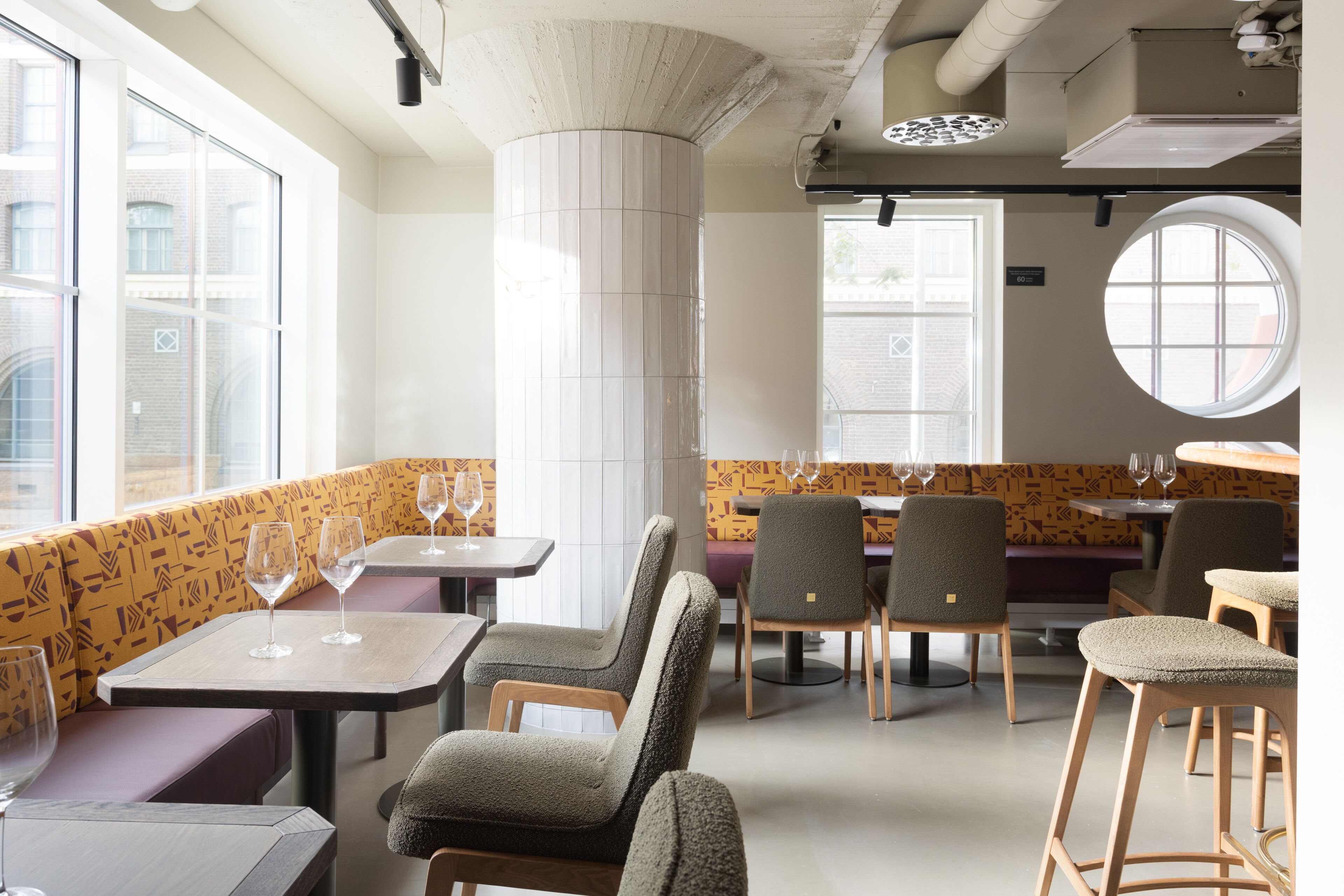 Interior de restaurante con mesas altas, asientos tipo banca acolchados y pared con diseño.