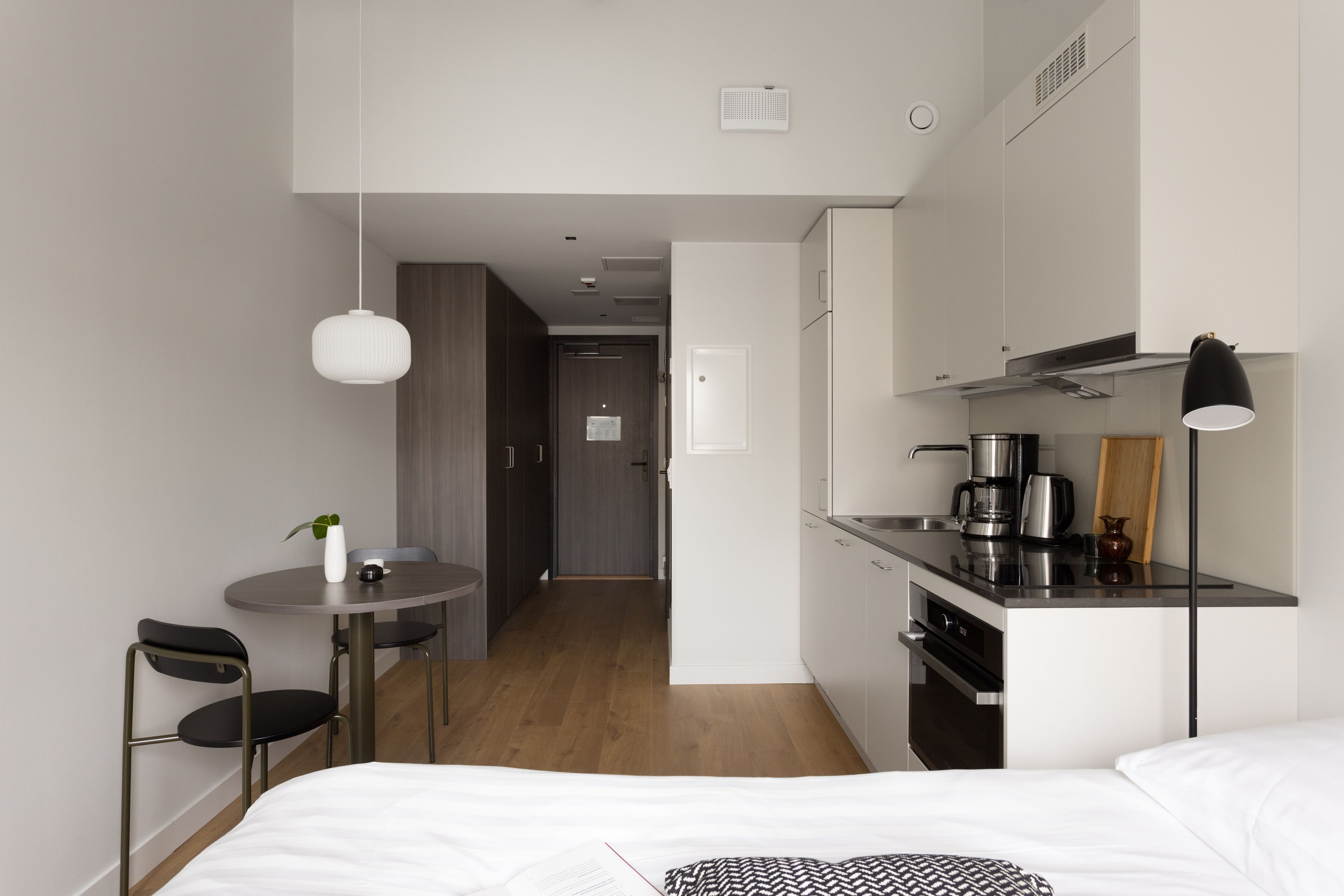 Habitación de hotel compacta con cama, una pequeña mesa de comedor con sillas, una kitchenette y un pasillo que conduce a otra habitación.
