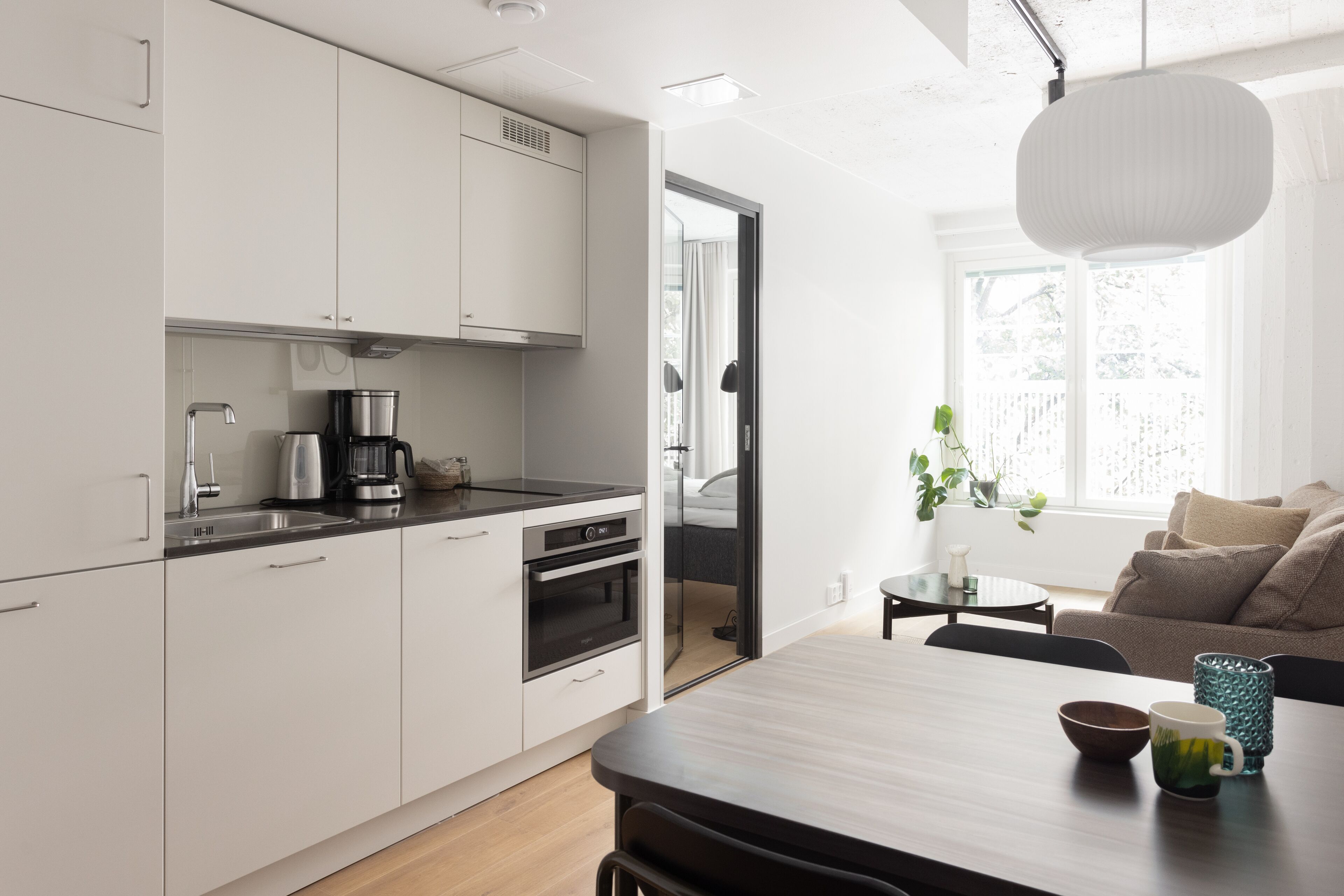 Une cuisine moderne avec des meubles blancs, une machine à café et un four. On y trouve un coin repas avec une table et des chaises, ainsi qu’une fenêtre donnant sur un paysage verdoyant.