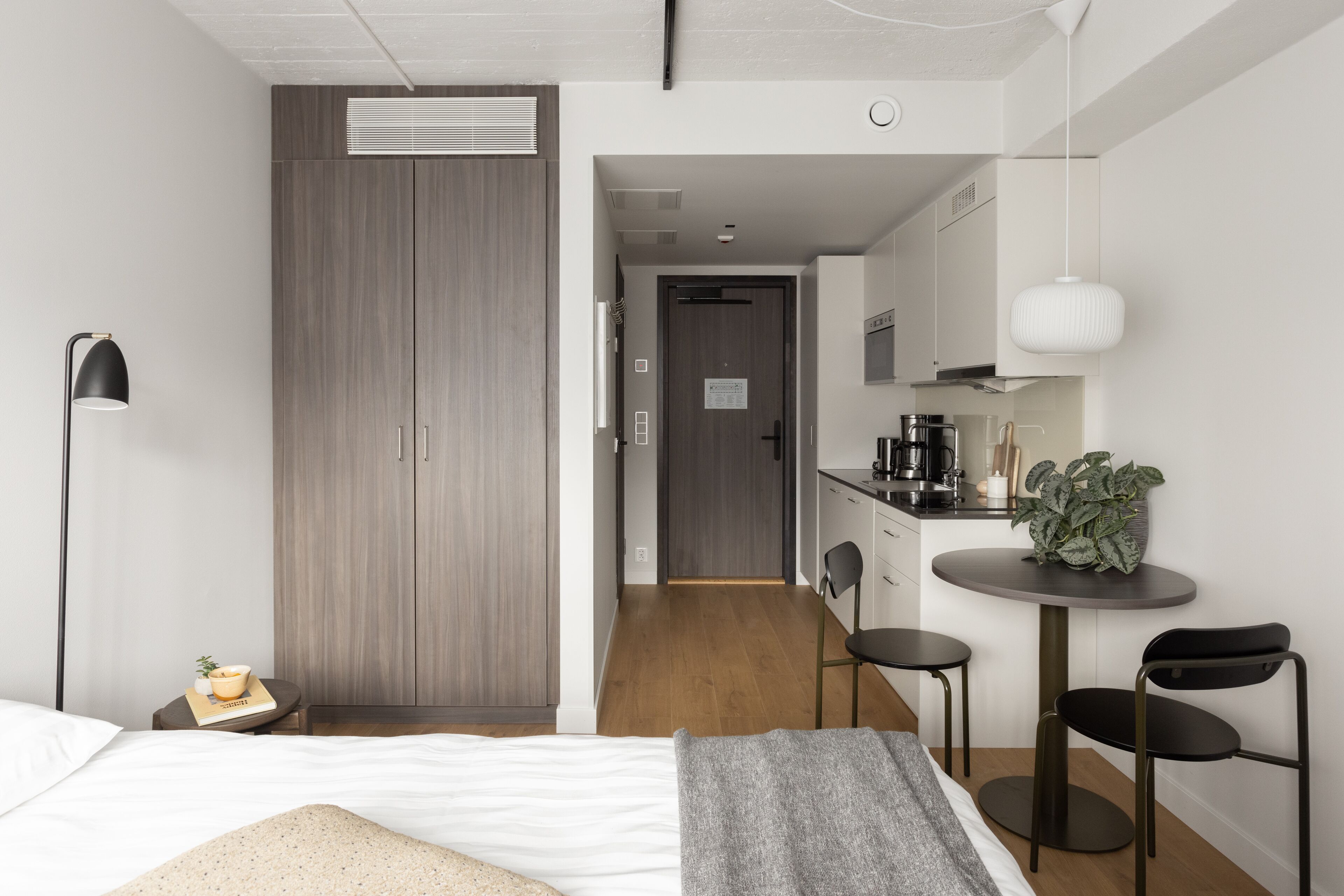A modern bedroom with a bed, bedside table, and a wardrobe. There is a kitchenette with a stove, sink, and cabinets. A small dining area with a table and chairs is visible.