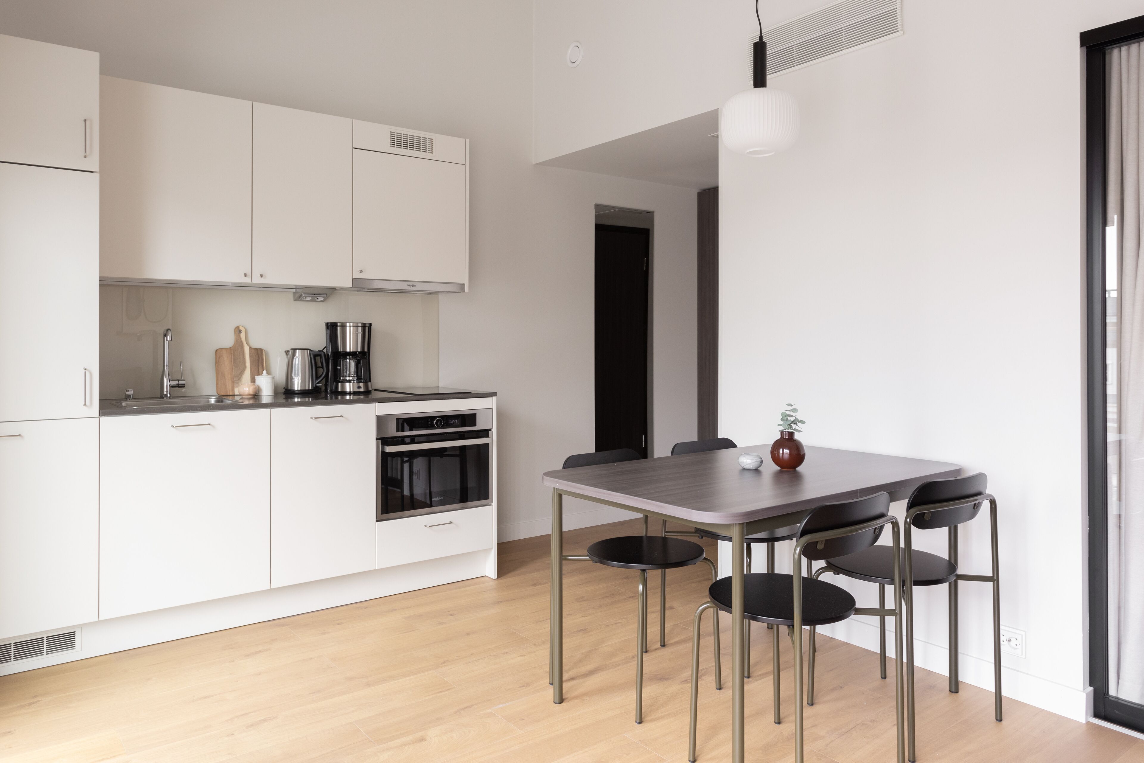 A modern kitchen with white cabinets, a built-in oven, a coffee maker, and a dining table with four chairs.