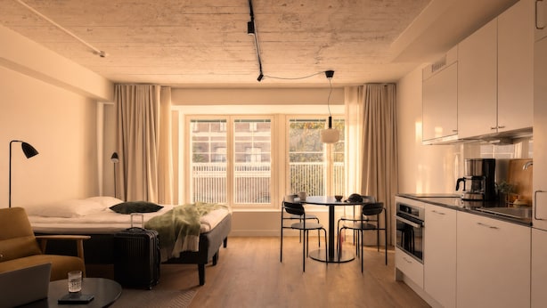 Ein kleines Hotelzimmer mit Bett, Schreibtisch mit Laptop, Koffer, Essbereich und Küchenzeile.