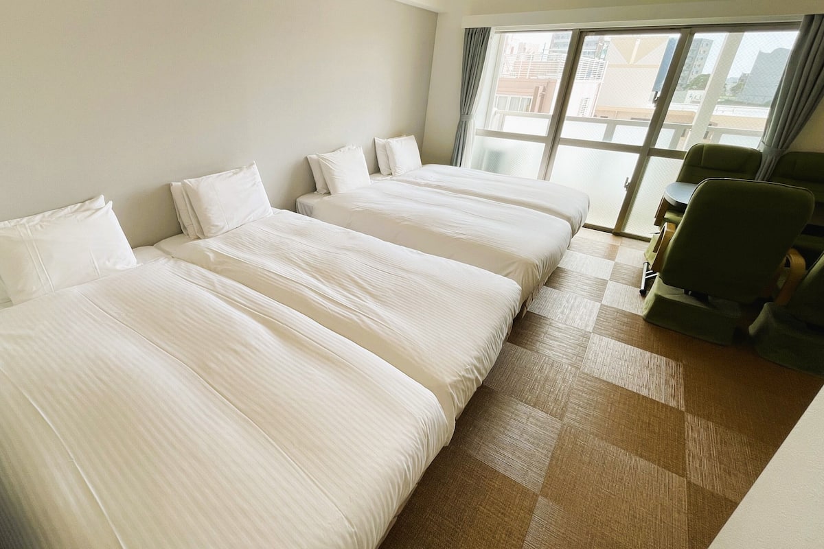 Quarto de hotel com três camas, uma janela com cortinas, uma mesa com cadeira e um balcão de recepção.