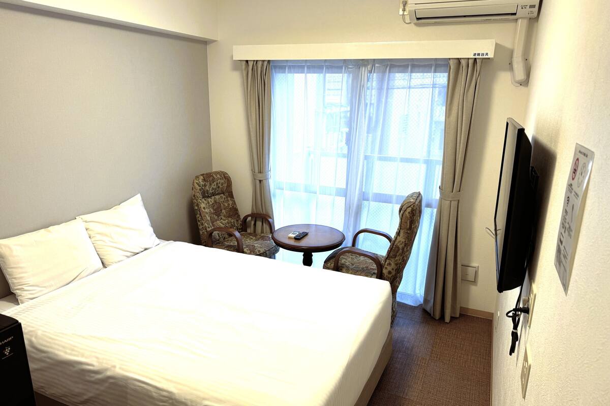 Quarto de hotel com uma cama grande, duas cadeiras, uma mesinha e uma televisão.