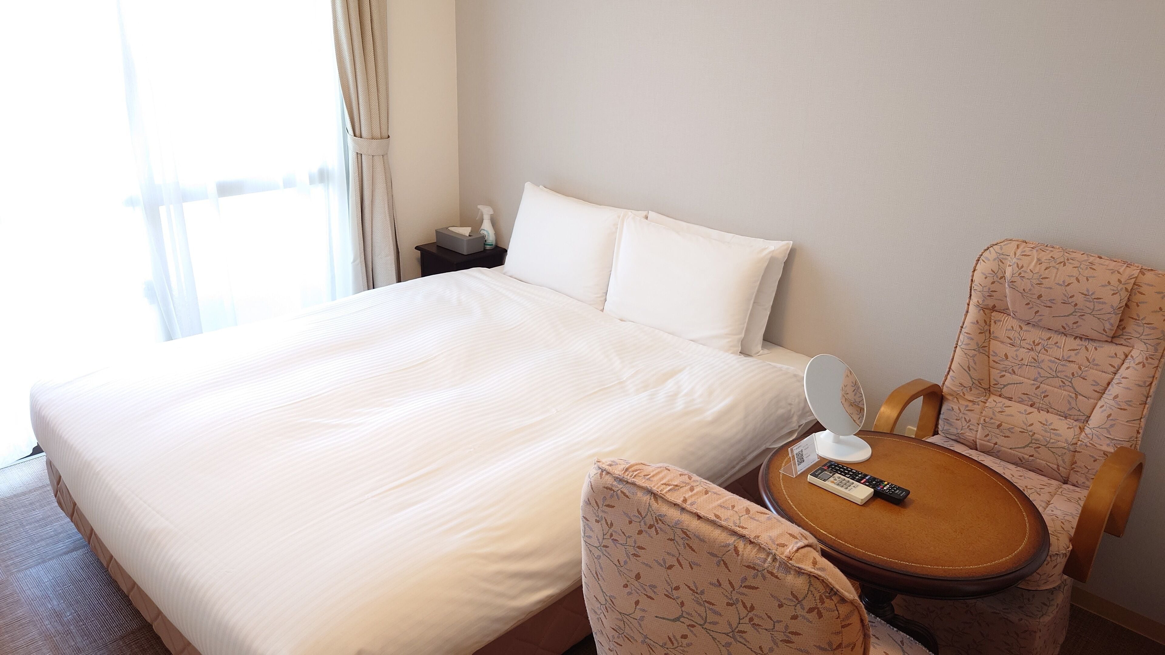 A hotel room with a large bed, a chair with a remote control, and a small table.