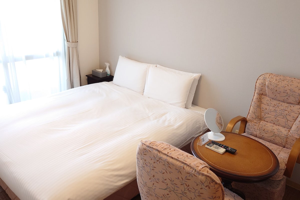 Quarto de hotel com uma cama grande, uma cadeira com controle remoto e uma mesinha.