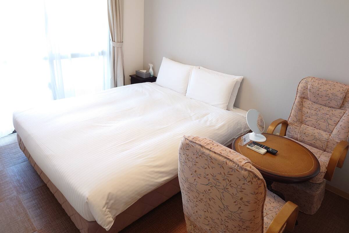 Quarto de hotel com uma cama grande, mesa de cabeceira com controle remoto, uma cadeira e janela com cortinas.