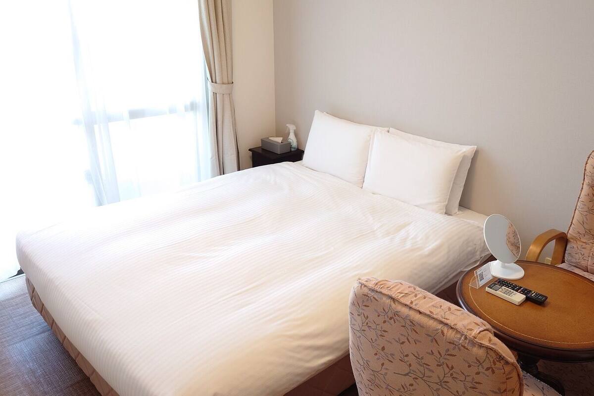 Quarto de hotel com uma cama grande, mesa de cabeceira com controle remoto, uma cadeira e janela com cortinas.