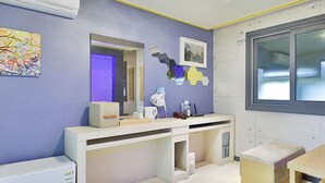 A modern bathroom with a white vanity, a mirror, and a sink. There is a window with blinds, a toilet, and a shower area. - The Apple Hotel (Busan)