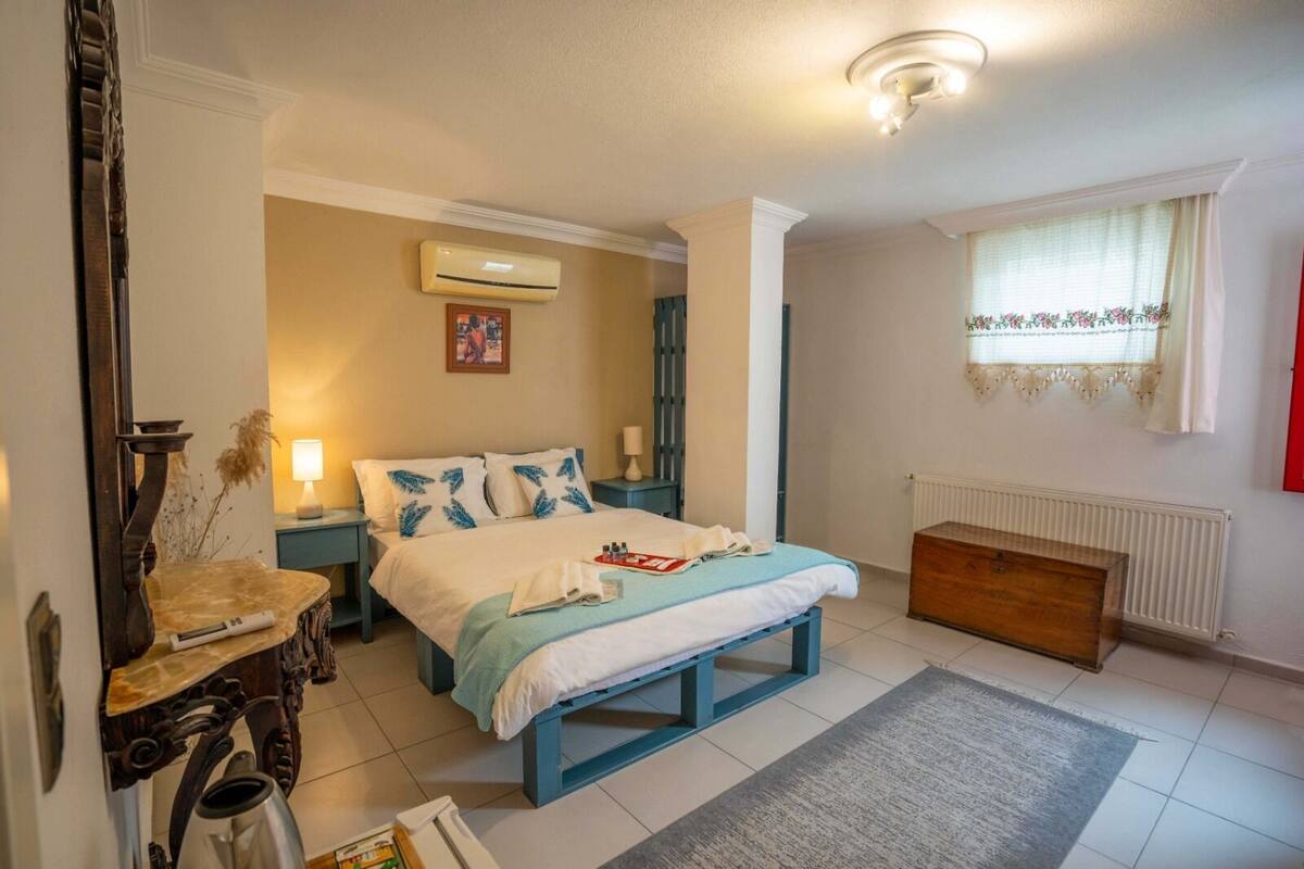 Quarto com cama, mesas de cabeceira, cômoda, radiador e janela com cortinas.