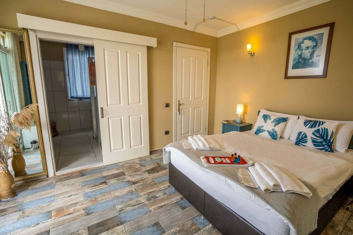 Quarto de hotel com uma cama grande, uma mesa de cabeceira com abajur, um espelho e uma porta que dá para outro quarto.