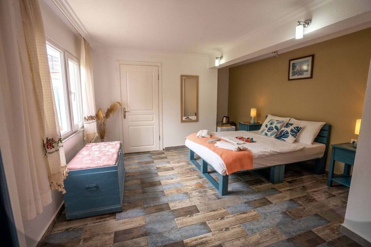 Quarto com cama, um banquinho, cômoda, espelho e janela com cortinas.