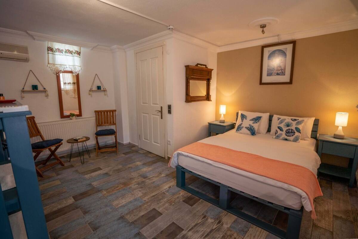 Quarto com cama, duas mesas de cabeceira, uma cadeira, uma mesinha, um espelho, uma porta e um radiador.