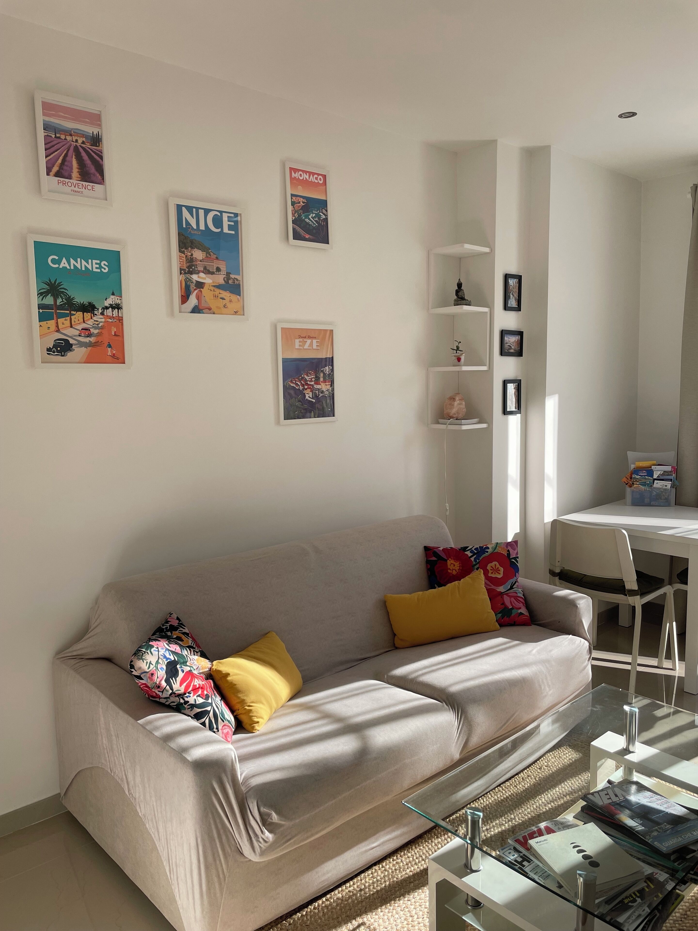 A living room with a beige sofa, colorful cushions, a glass coffee table, and a white TV stand. There are posters on the wall and a small white shelf with decorative items.