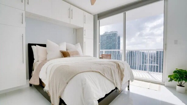 A modern bedroom with a large bed, white walls, and a view of a cityscape through a large window. - Chic Penthouse: 3BR | Pool | Gym| 2 Parking Spots (Miami)