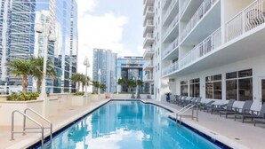 A swimming pool with a sun deck, surrounded by high-rise buildings and palm trees. - Chic Penthouse: 3BR | Pool | Gym| 2 Parking Spots (Miami)