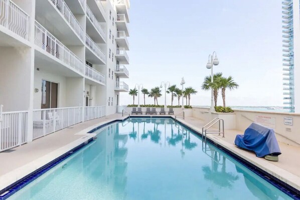 A swimming pool with a clear view of the ocean, surrounded by a white building with balconies and palm trees.