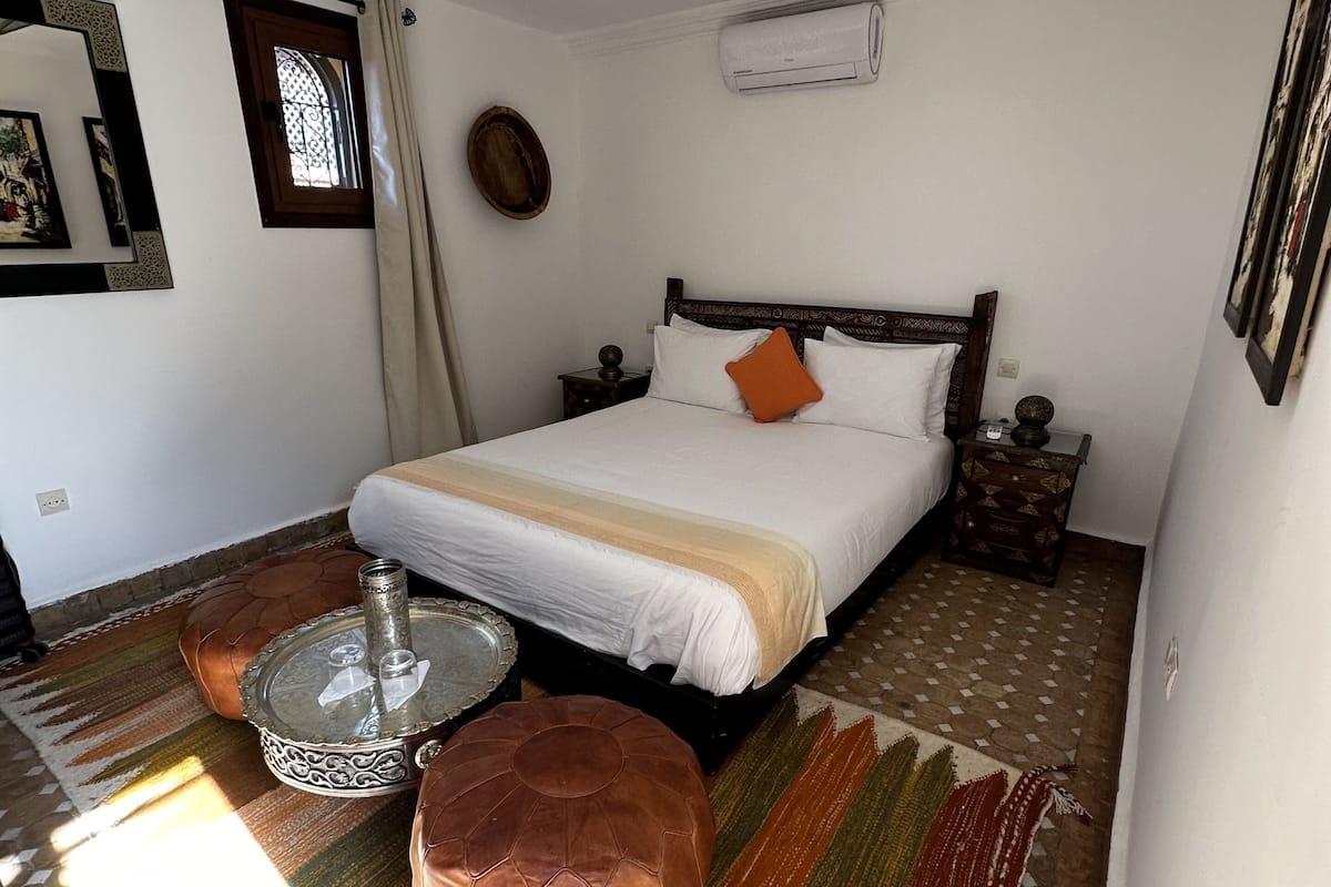 A bedroom with a bed, a nightstand, a decorative lamp, a window with curtains, and a patterned rug.