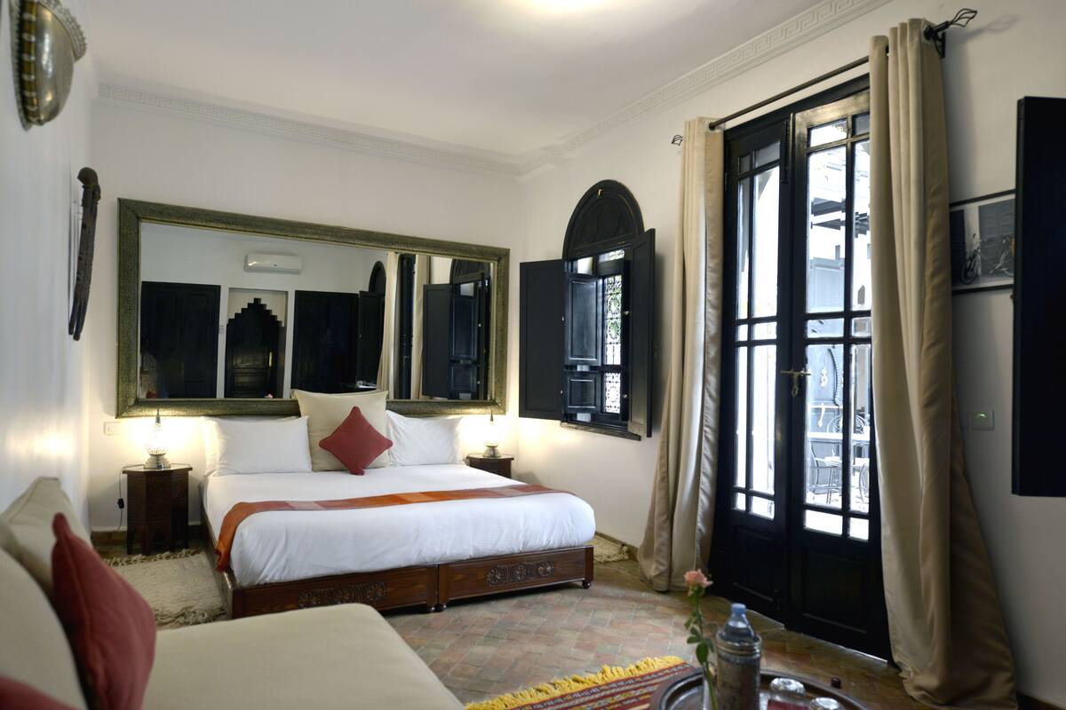 A bedroom with a large bed, a nightstand, a mirror, and a window with curtains.