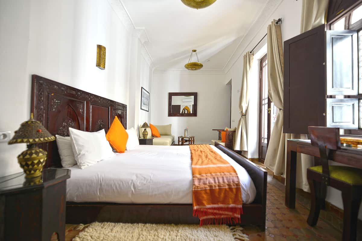 A neatly arranged bedroom with a large bed, a bedside table with a lamp, a chair, and a desk.