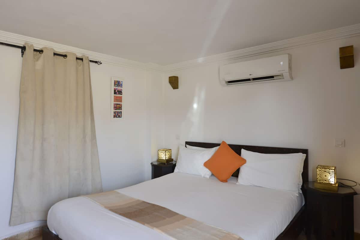 A neatly made bed with white and orange pillows, a beige blanket, and a wooden headboard. There is a small table with a lamp and a picture frame beside the bed. An air conditioning unit is mounted on the wall above the bed.