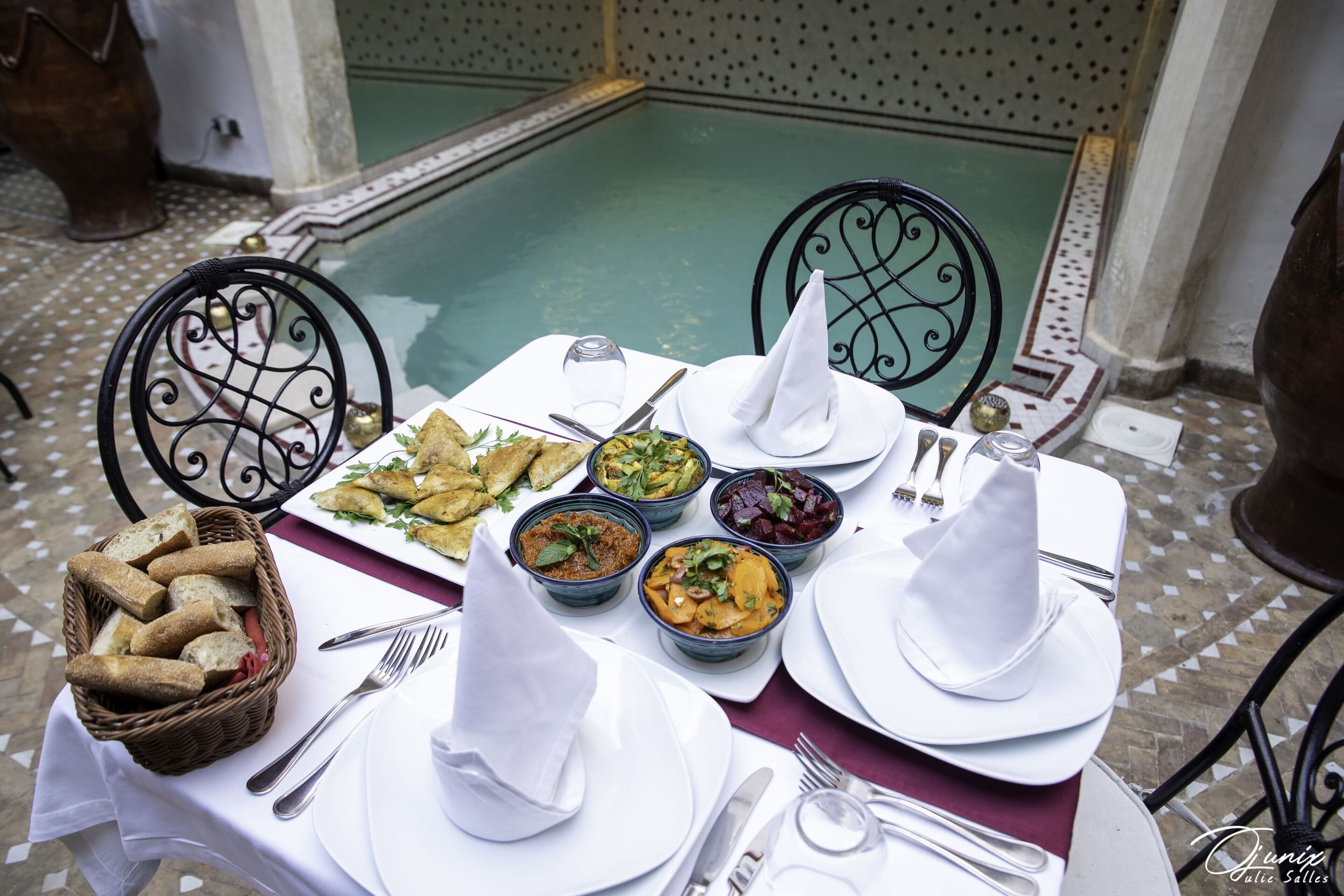 A table set for a meal with various dishes, including bread, salads, and bowls of food, in an outdoor dining area with a pool in the background.