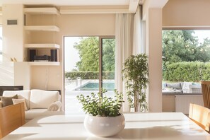A modern living room with a white sofa, a dining table with a white vase, and a large window with a view of a swimming pool and greenery.
