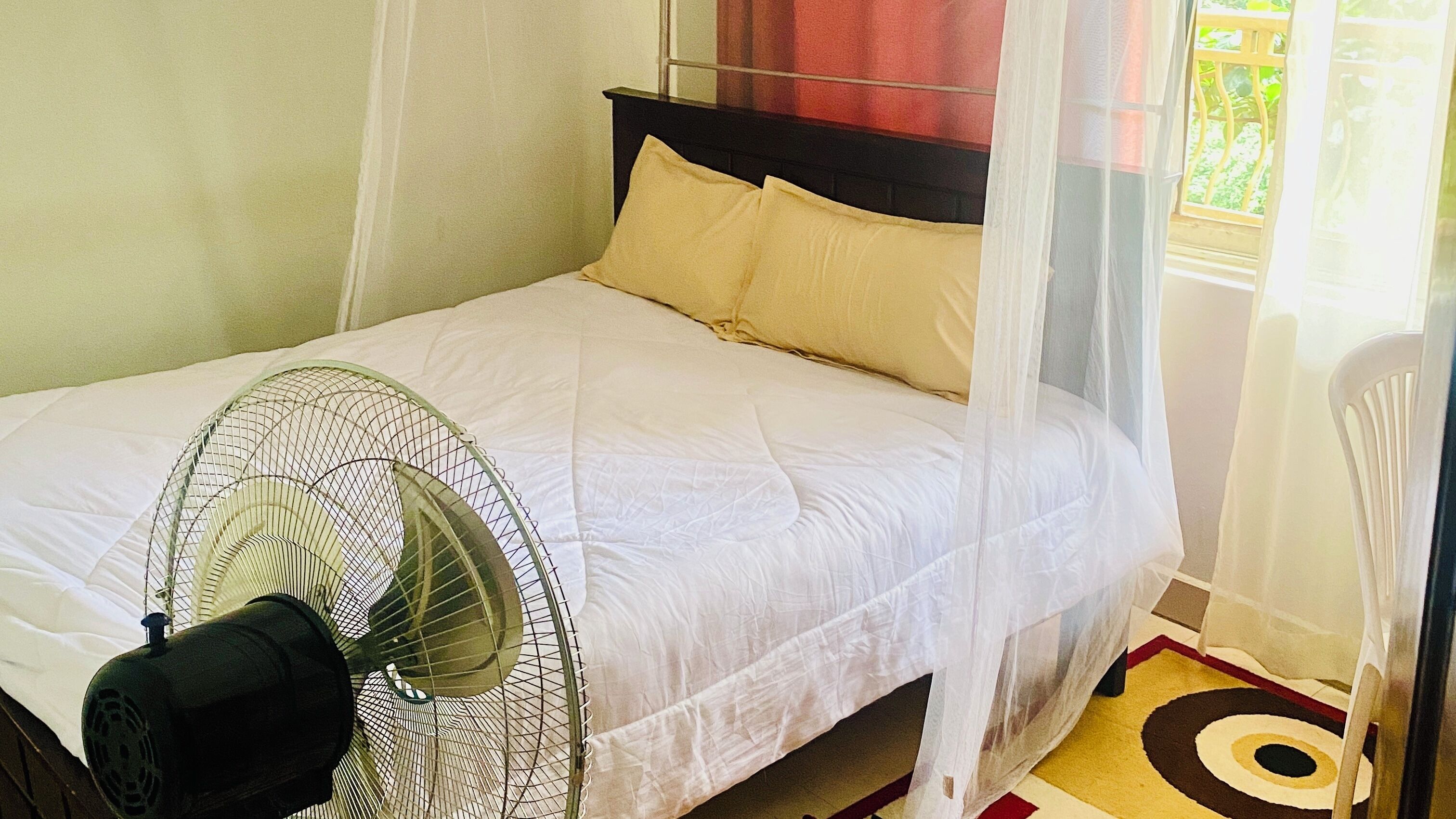 A bed with a white bedspread and two pillows, a standing fan, and a chair with a white cushion.