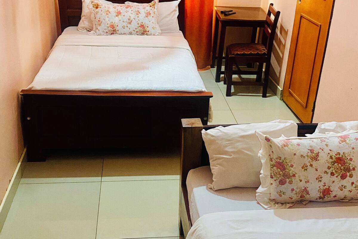 A hotel room with two beds, each with floral-patterned pillows, a wooden nightstand, a small table with a chair, and a wooden door.