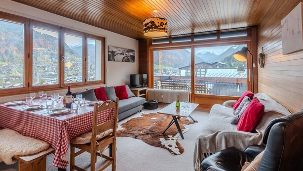 A cozy room with a wooden ceiling, large windows offering mountain views, a dining table set with glasses and plates, and a sofa with red cushions. - 3-room apartment 5 min from Morzine center (Morzine)
