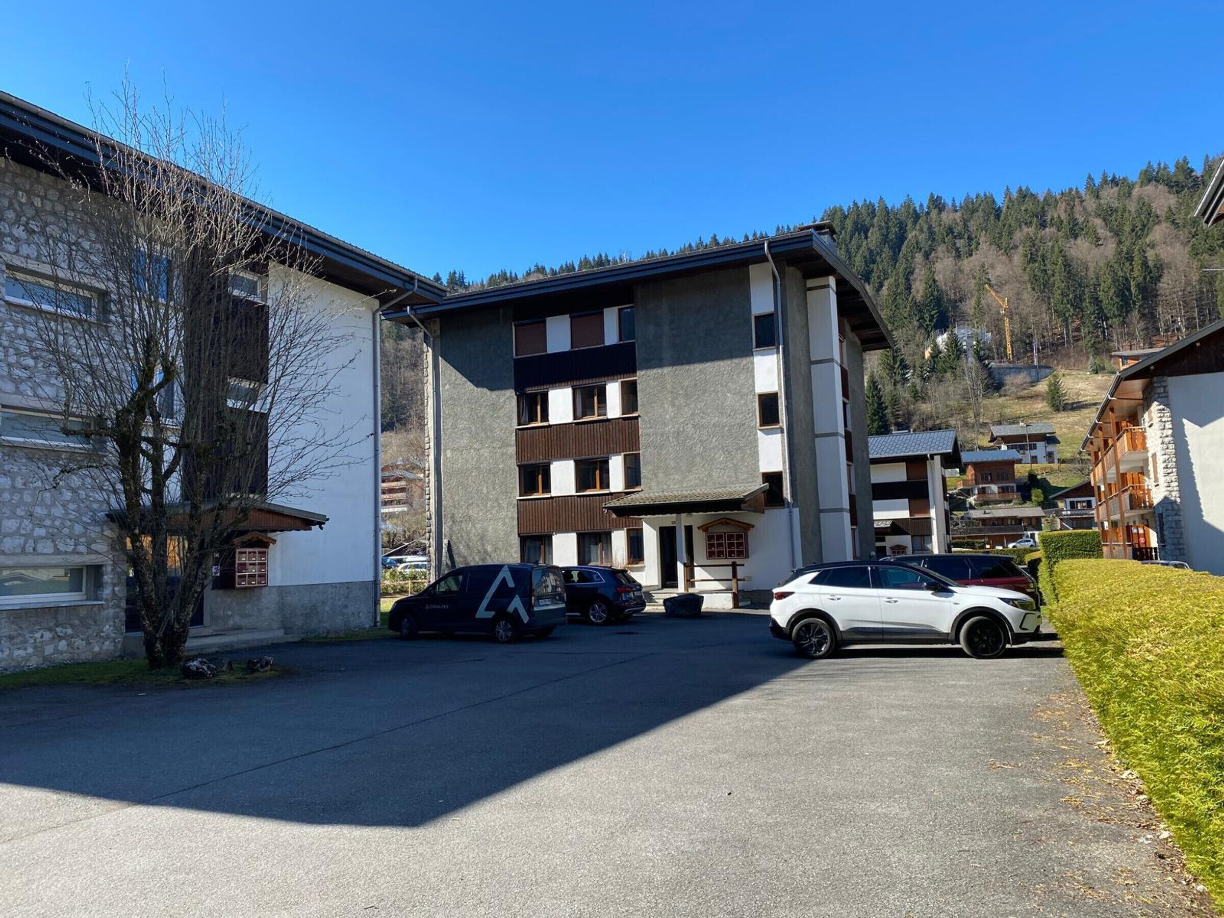 Un immeuble à plusieurs étages, doté d’une façade en pierre et d’un toit plat. Plusieurs voitures sont garées devant, et une zone herbeuse se trouve sur la droite.