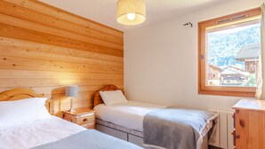 A room with wooden panel walls, a bed with a headboard, a bedside table with a lamp, and a window with a view of buildings. - Comfortable 4-room in Morzine, terrace, garage, near center (Morzine)