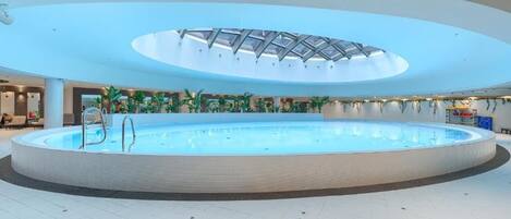 A large indoor swimming pool with a glass ceiling, surrounded by a white tiled floor and indoor plants.