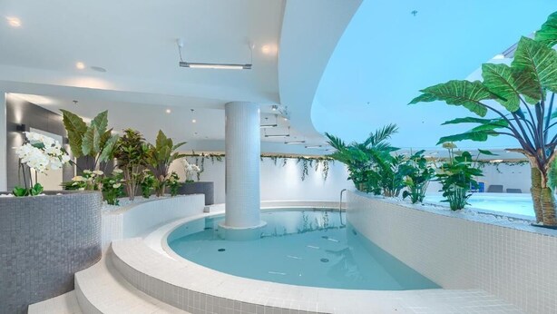 A modern indoor pool area with a curved design, surrounded by various plants and a large column.