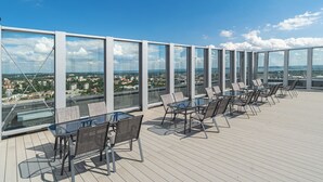 A rooftop terrace with glass walls, outdoor seating, and a view of the cityscape.