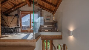 A spacious room with a high ceiling and wooden beams, a cozy seating area with a sofa and chairs, a staircase leading to an upper level, and a view of mountains through large windows. - Charming renovated chalet in Morzine center with parking, sleeps 4. (Morzine)