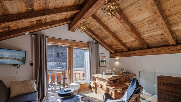 A cozy room with a wooden ceiling, a sofa, a chair, a dining table with chairs, and a view of a mountain landscape through a large window. - Charming renovated chalet in Morzine center with parking, sleeps 4. (Morzine)