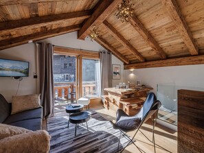 A cozy room with a wooden ceiling, a sofa, a chair, a dining table with chairs, and a view of a mountain landscape through a large window.