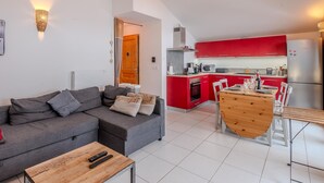 A modern apartment with a grey sofa, a wooden coffee table, a dining table with chairs, a kitchen with red cabinets, and a wooden door.