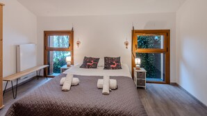 A bedroom with a large bed, two bedside tables, a bench, and two windows with a view of greenery.