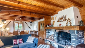 A cozy cabin interior with a stone fireplace, wooden beams, a dining area, and a blue sofa.