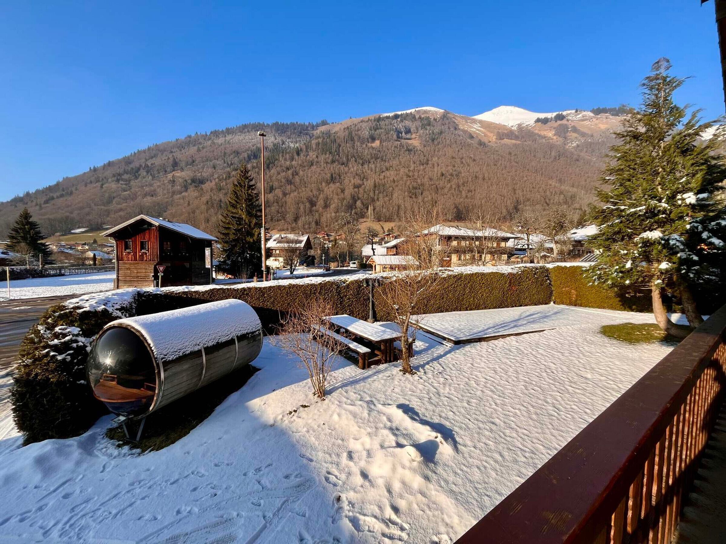 Ein verschneiter Außenbereich mit einer Holzterrasse, einer zylindrischen Struktur und einem Blick auf einen Bergkamm im Hintergrund.
